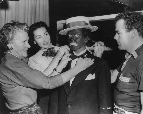 1951:  American singer and actor Doris Day frowns while a makeup man touches up the blackface makeup on her ear and wardrobe women fix her tie, on the set of director Michael Curtiz's film, 'I'll See You in My Dreams'. Day wears a straw boater and a tuxedo.  (Photo by Hulton Archive/Getty Images)