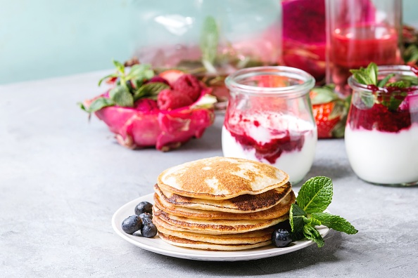 Stack of homemade pancakes served on plate with berries, mint, glass jars of yogurt, bottle of lemonade, fruit salad in pink dragon fruit over grey texture table