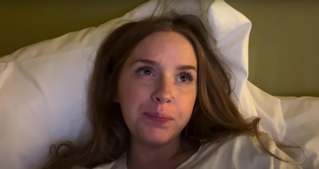 Closeup shot of Ashlyn Vanhorn's face and shoulders while she lies back on a bed with white sheets and looks up and to the side