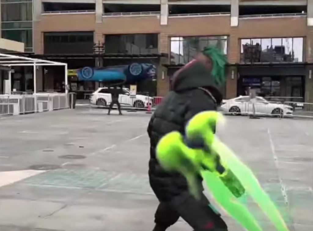 Virtual rapper FN Meka adopts a fighting position in an outdoor parking lot, wielding a green fiery blade as a man in the distance holds a car above his head