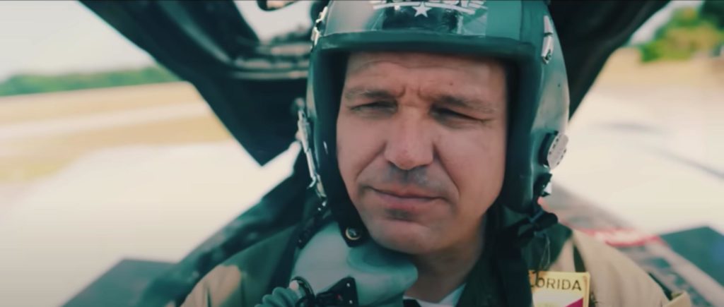 Ron DeSantis in the cockpit of a military-style plane, wearing a pilot's helmet and squinting into the sun