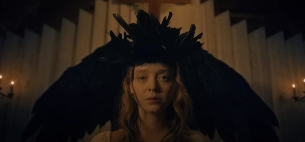 Young woman wearing black feather headdress stares at the camera in a dimly lit room