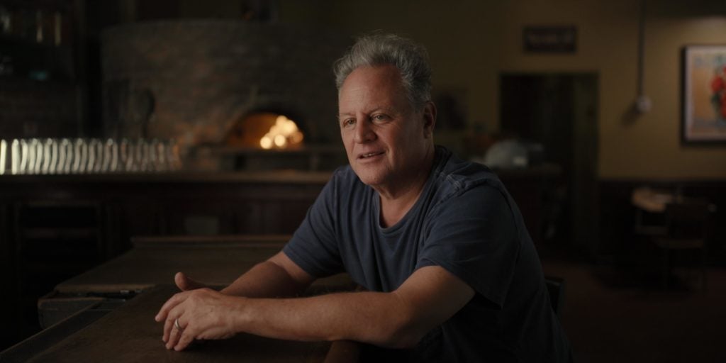 Chris Bianco rests his arms on a table in front of a wood fire pizza oven in Chef's Table: Pizza on Netflix