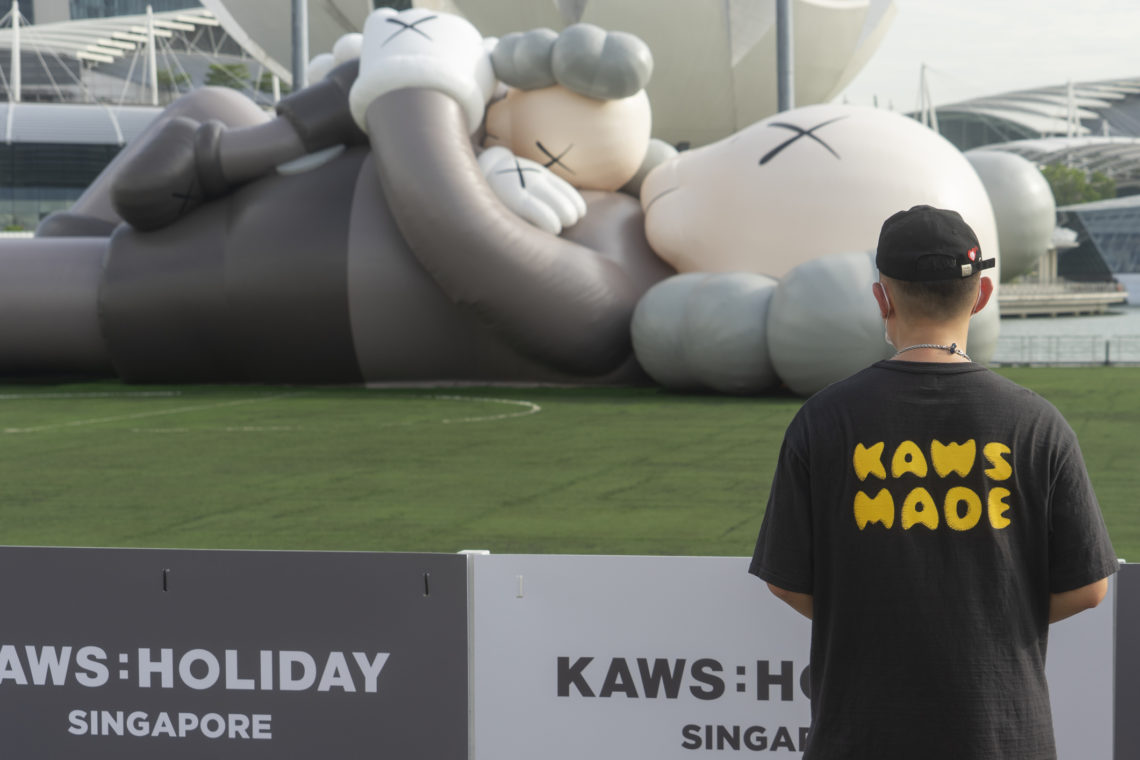 Large sculpture of two figures laying down, by Kaws, as exhibited in Singapore, with a man wearing a T-shirt with the words "KAWS MADE" in the foreground