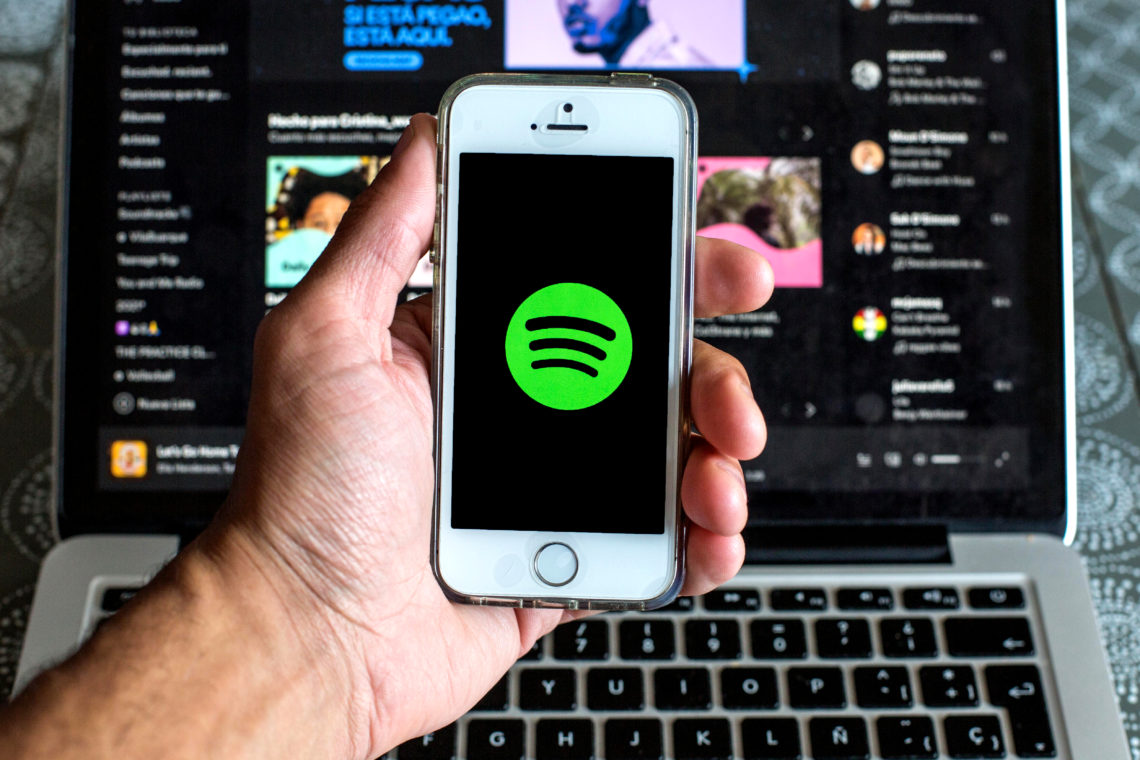 A left hand holds a smart phone with the Spotify logo on it screen, in front of a laptop screen with Spotify open