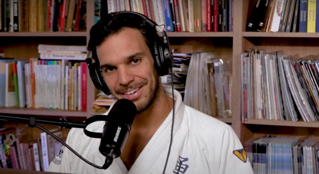 Jiu-jitsu instructor Joaquim Valente wears headphones and speaks into a mic while recording a podcast, wearing jiu-jitsu clothing and sitting in front of a bookcase