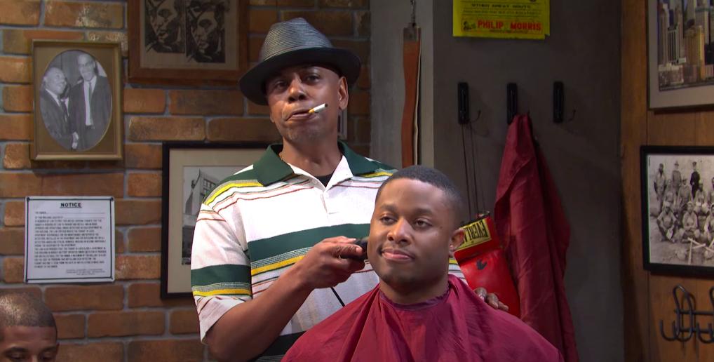 Dave Chappelle as a barber trims a client's hair while smoking a cigarette in an SNL skit
