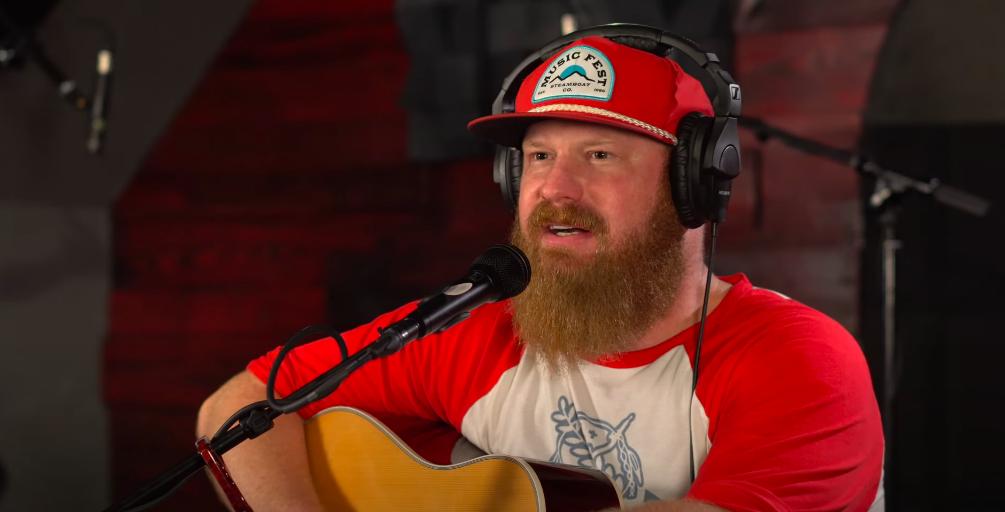 Jake Flint wearing a red cap and black headphones speaks into a microphone with an acoustic guitar on his lap