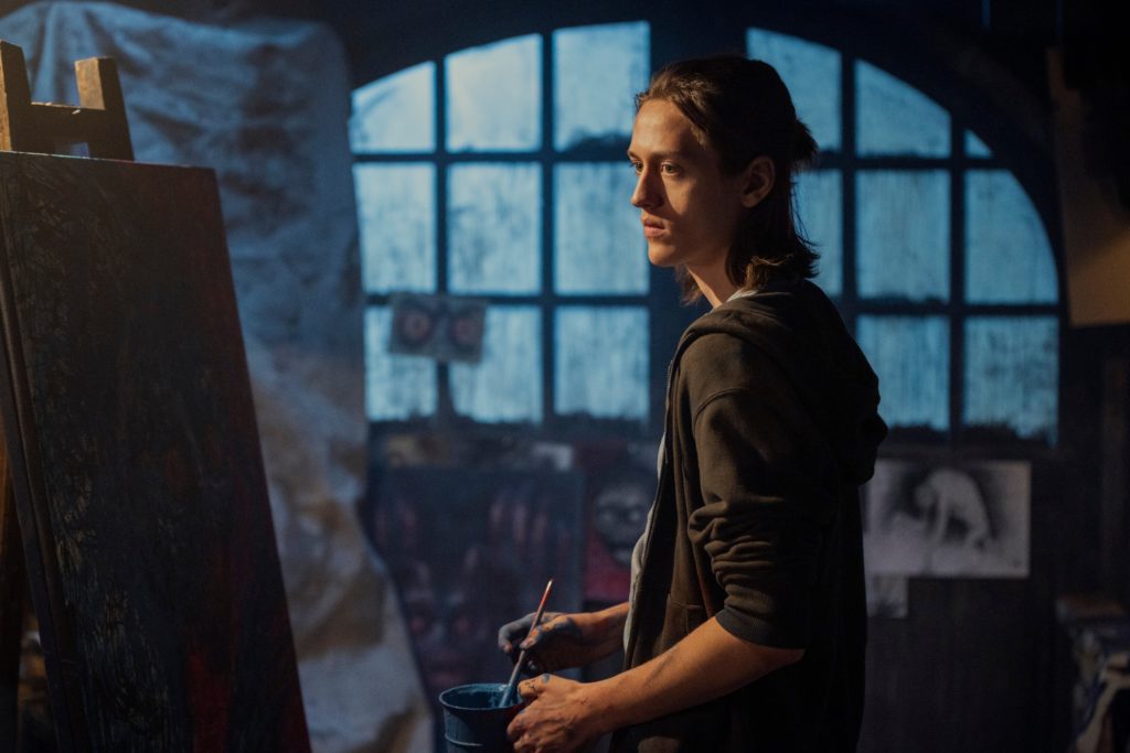 Xavier stands in front of a canvas with a paintbrush in his hand in Wednesday episode 6
