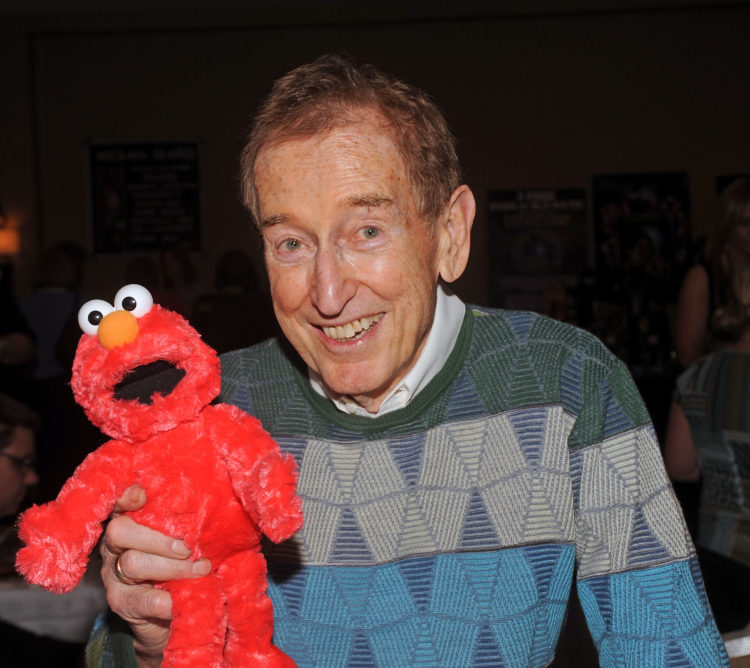 Bob McGrath holds Elmo in his right hand, wearing a grey sweater and smiling at the camera