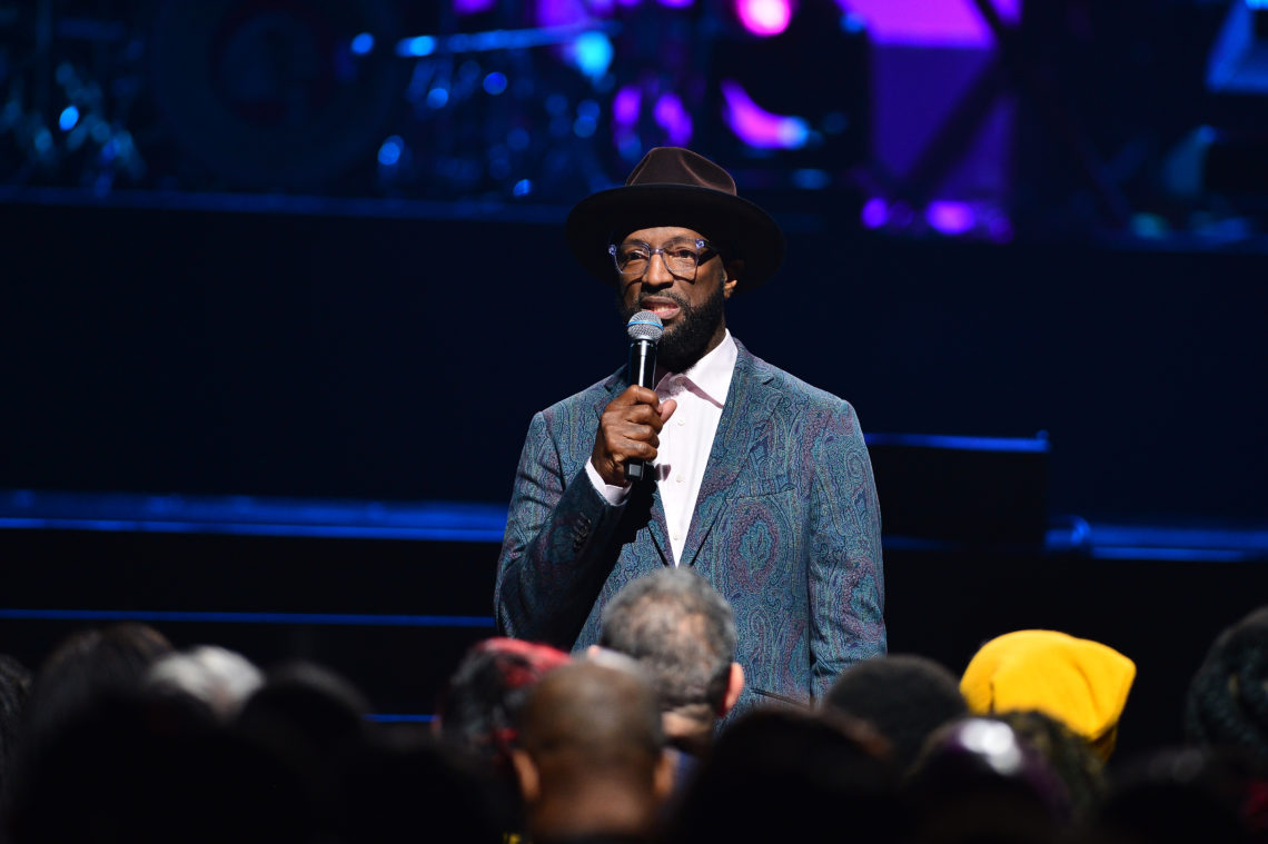 Rickey Smiley hosts the 21st Annual Super Bowl Gospel Celebration at the James L. Knight Center in 2020, holding a microphone to his mouth and wearing a stylish suit
