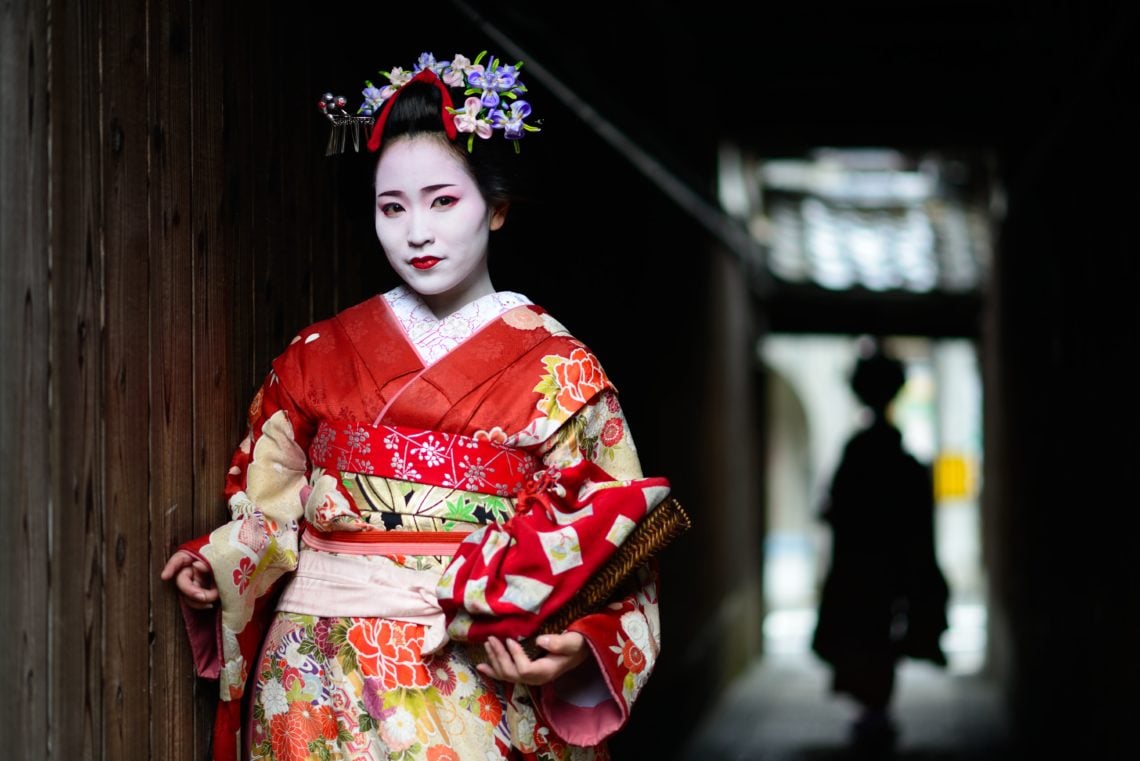 A Geisha woman with flowers in her hair stands in a dark corridor looking at the camera.