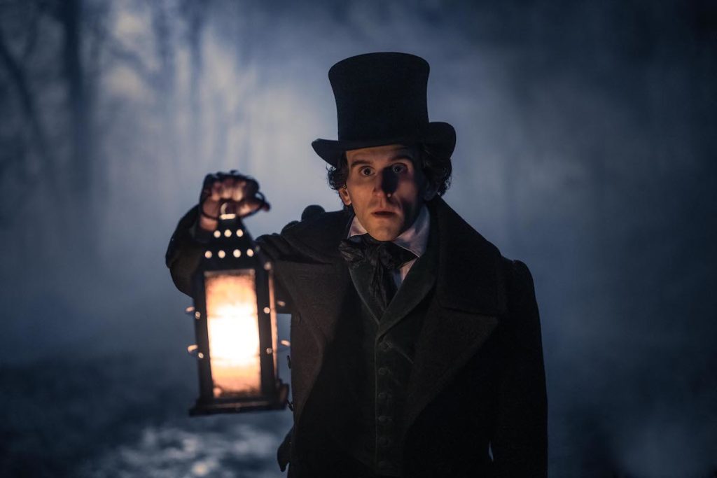 Harry Melling as Edgar Allen Poe holds a lamp light in a dark misty woods in The Pale Blue Eye
